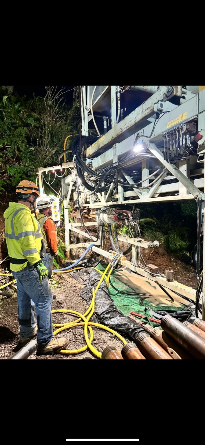 Crew monitoring geotechnical drill with hydraulic hoses and lights at night